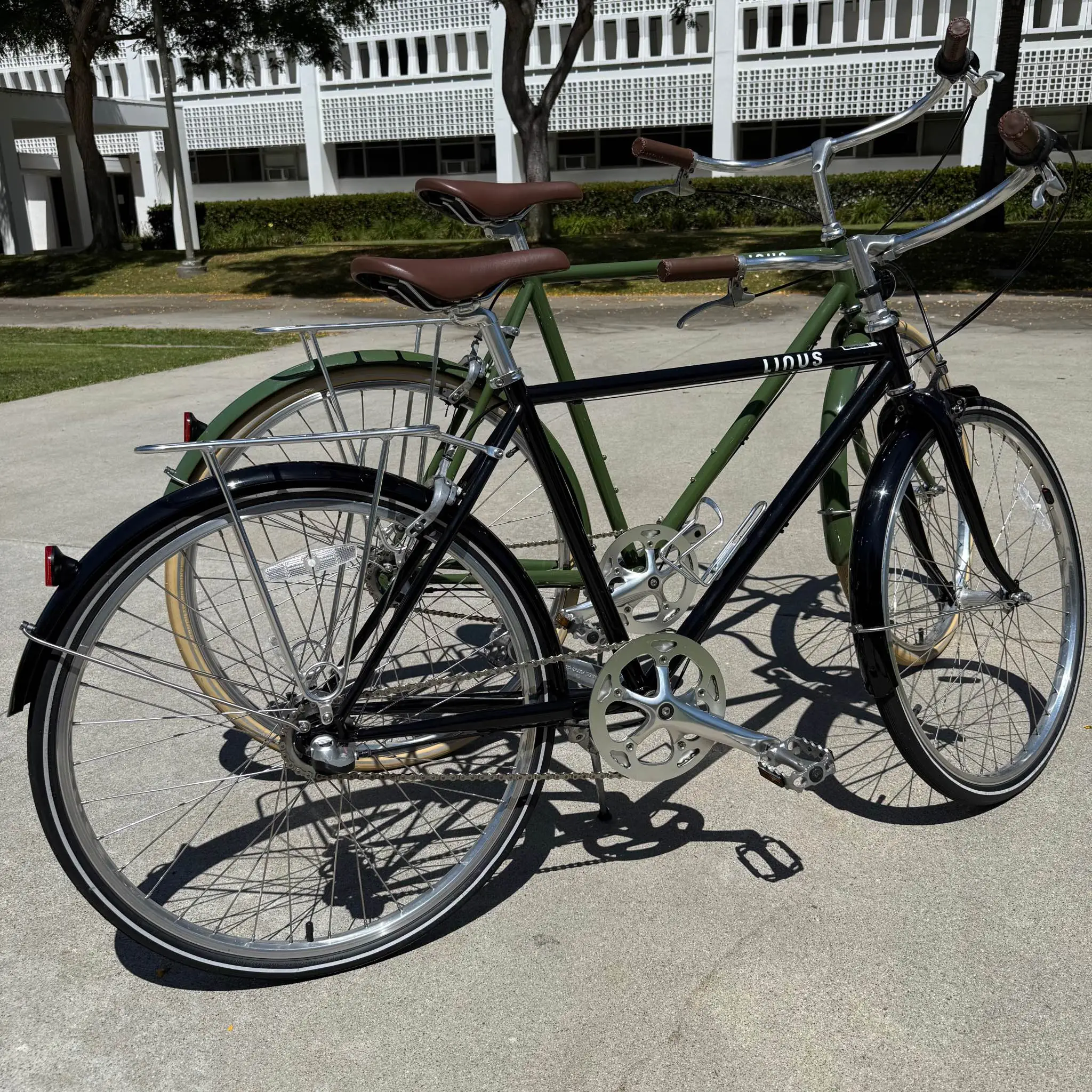Two restored bikes (an olive green mixte and a black step-through) parked side by side outside a modernist building.